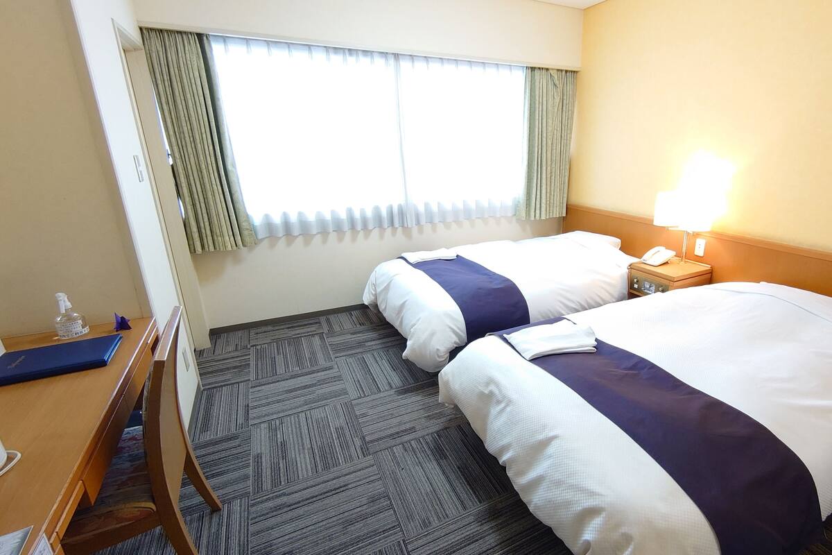 A hotel room with two beds, a desk, a chair, a lamp, and a window with curtains.