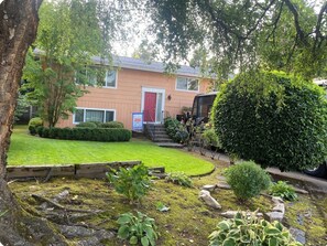 A residential building with a red door, a well-maintained lawn, and a variety of shrubs and plants. - Extended Stay • Walk to Restaurants Cafés & Food Carts  • Patio Large Yard • Pet (West Linn)