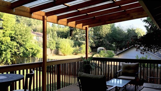 Une terrasse en bois agrémentée d’une pergola, de mobilier d’extérieur et offrant une vue sur un jardin avec des arbres et une maison.