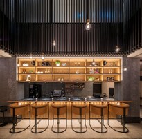 Zona de bar moderna con una barra alargada, varios taburetes y estanterías con distintos objetos.