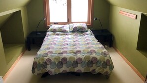 Une petite chambre avec un lit simple, un plafond en pente, une fenêtre avec des cadres en bois, deux tables de chevet avec des lampes et une couvre-lit aux couleurs vives.