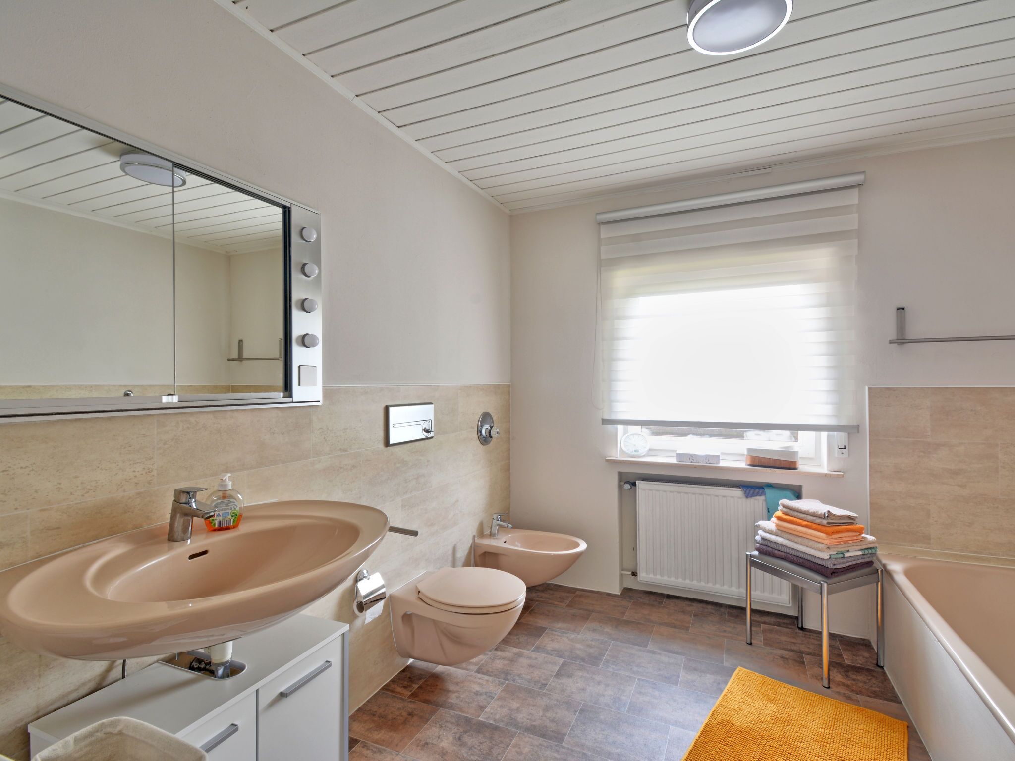 Et moderne badeværelse med en stor vask, toilet og bidet. Der er et spejl, et vindue med persienner og en radiator.