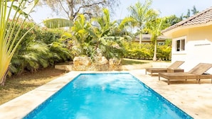 Una piscina rectangular con agua azul cristalina, rodeada por un muro de piedra y exuberante vegetación tropical. A la derecha de la piscina hay dos sillones de mimbre.