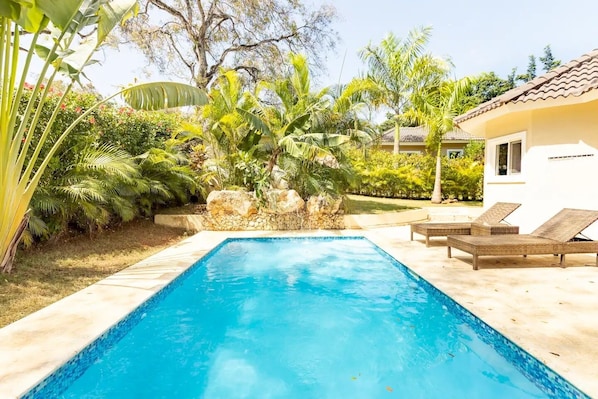 A rectangular swimming pool with clear blue water, surrounded by a stone wall and lush tropical plants. There are two wicker lounge chairs on the right side of the pool. - Private Pool & Jacuzzi Villa in Sosúa (Sosúa)