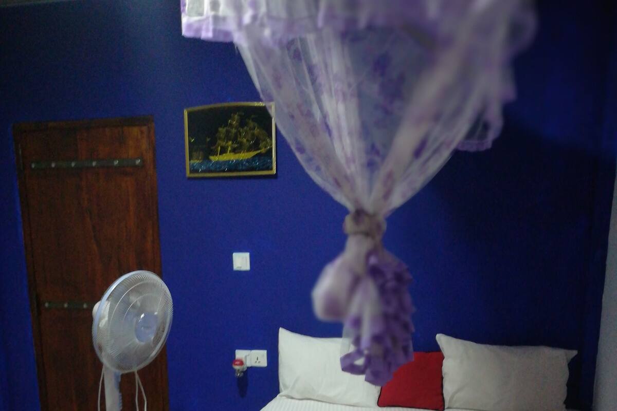 Quarto com cama, ventoinha de cabeceira, porta de madeira e parede com um quadro.