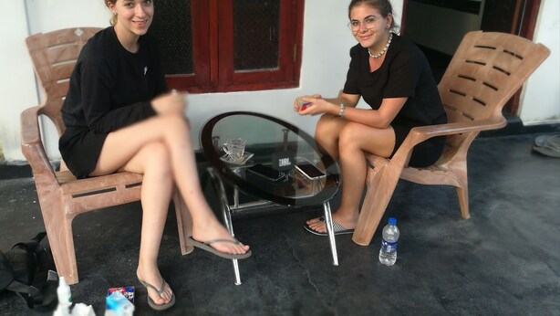 Two people sitting on plastic chairs outside a building with a window and a door in the background. - Tiny Holiday Resort (Dambulla)