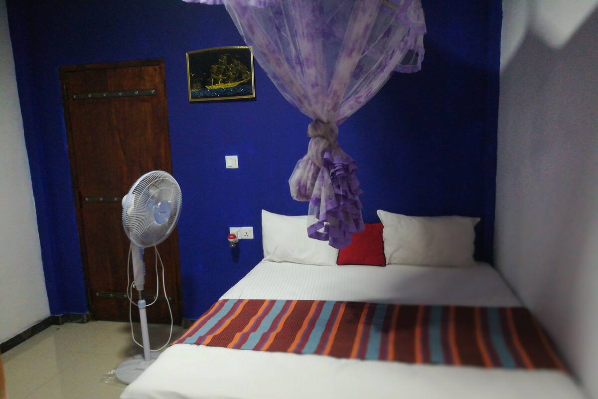 Quarto com cama, ventilador, porta de madeira e tomada na parede.