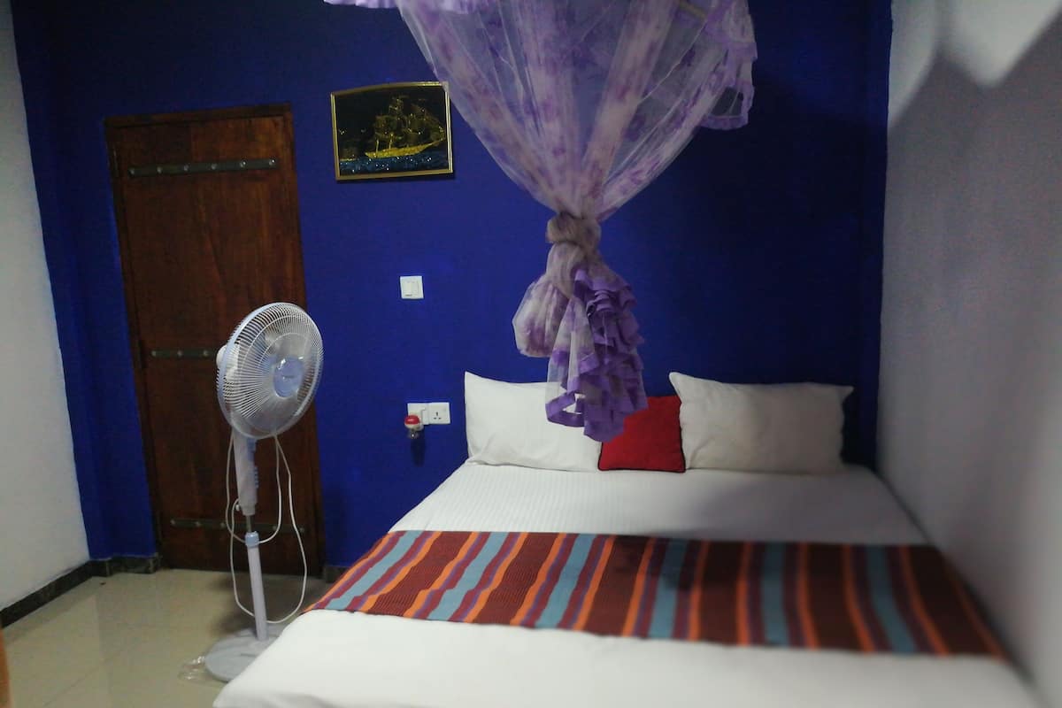 Quarto com cama, ventilador, dossel roxo, porta de madeira e interruptor de luz fixado na parede.