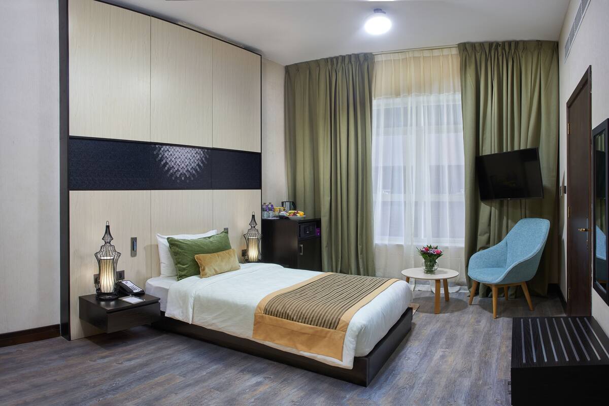 Une chambre d’hôtel comprenant un lit, une table de chevet avec une lampe, une petite table avec un vase, une chaise, un téléviseur et une armoire.