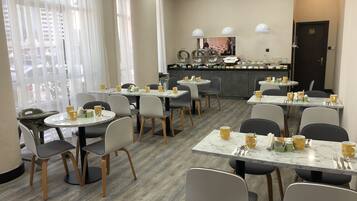 A hotel dining area with round tables, chairs, and yellow mugs. There are large windows with sheer curtains, a kitchen area in the background, and a door with a number sign.