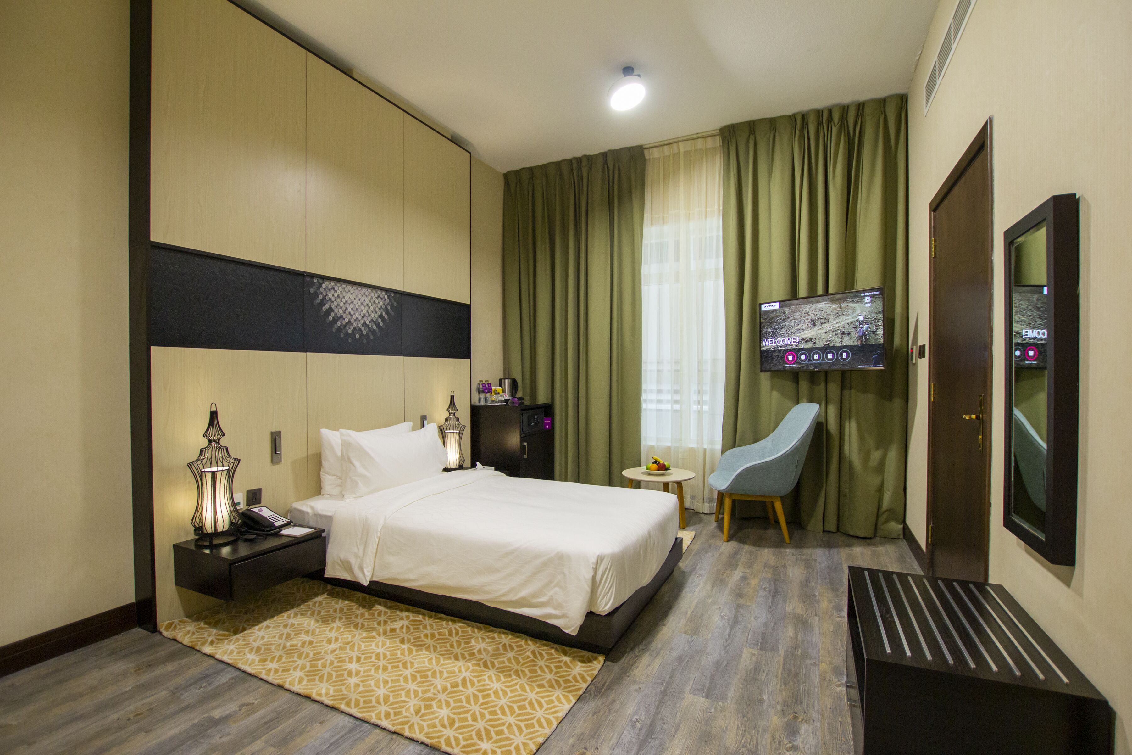 A hotel room with a bed, bedside table, lamp, TV, chair, and a small table with fruit.
