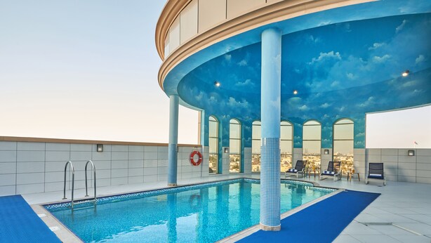 An indoor swimming pool with a blue-tiled floor, a lifebuoy, and a ceiling painted to resemble a sky with clouds. - Express By TIME - Al Khan (Sharjah)
