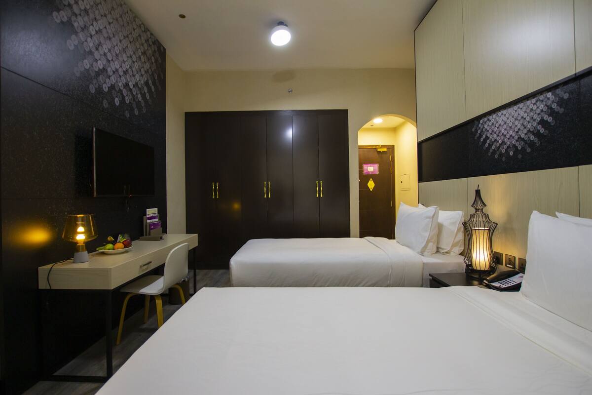 Une chambre d’hôtel équipée d’un bureau, de deux lits, d’une armoire et d’une table de chevet avec une lampe.