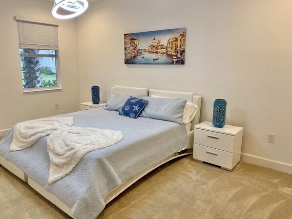 Een slaapkamer met een bed, witte nachtkastjes, een schilderij van een gracht en een raam met jaloezieën.