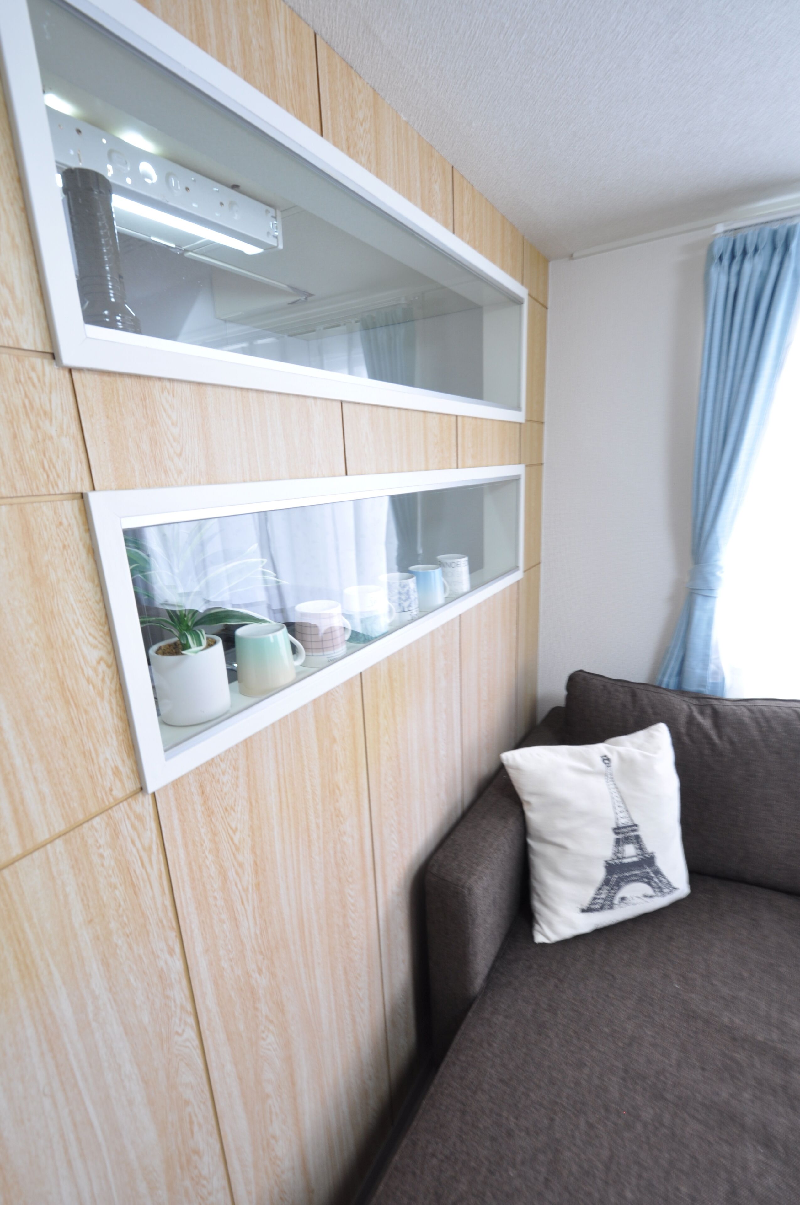 Une pièce moderne avec un mur en bois, une étagère intégrée ornée d’objets décoratifs, un canapé gris avec un coussin et une fenêtre avec des rideaux bleus.