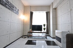 A hotel room with a flat-screen TV on a wooden stand, a small table with a potted plant, a wall-mounted heater, and a large window with curtains. - AMS Hiragishi 11 502 (Sapporo)