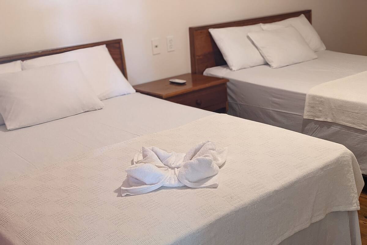 Two single beds with white linens and towels folded on them, a wooden nightstand with a remote control, and a wooden dresser in the background.