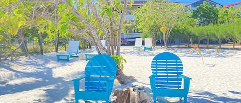 Zona de playa con dos sillas azules y una mesa de madera, rodeada de árboles y un edificio al fondo.