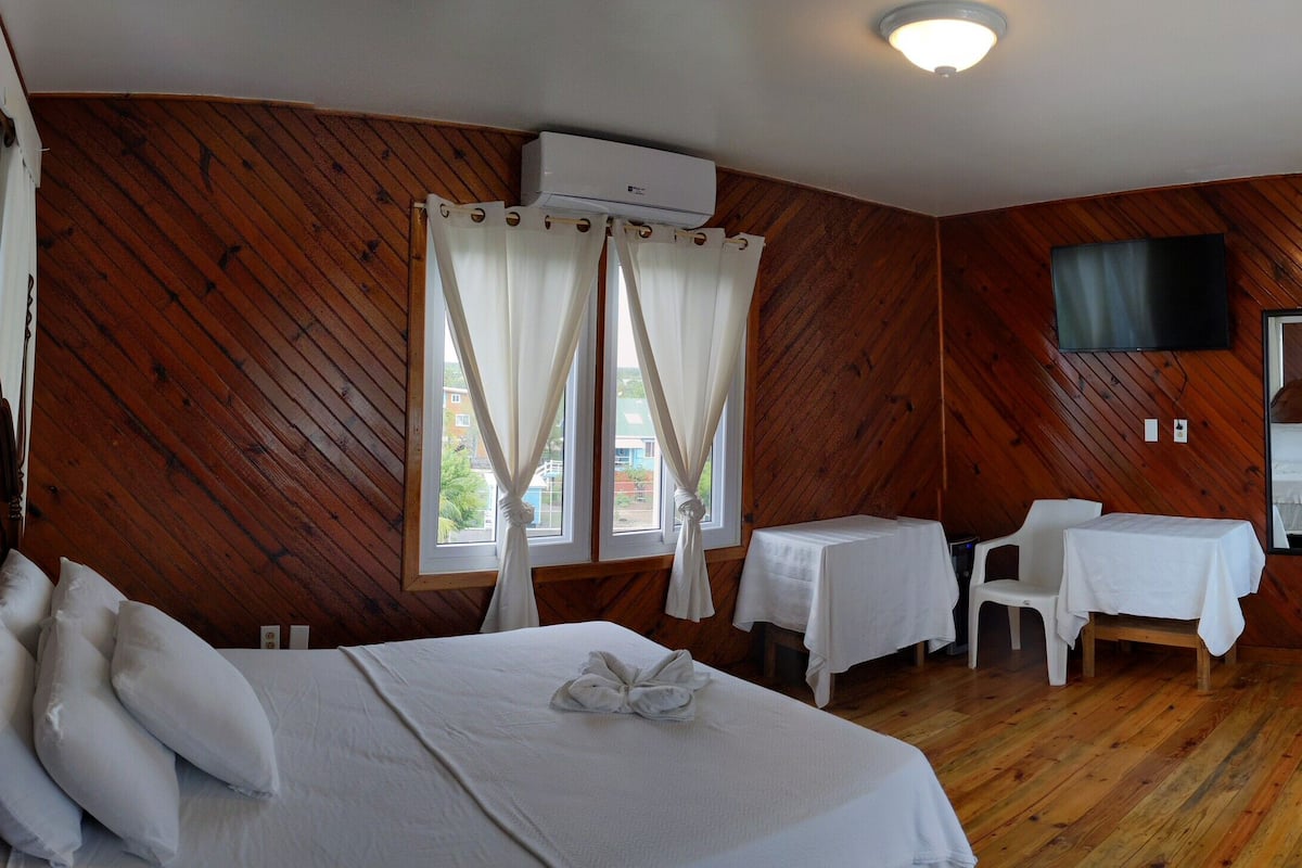 A wooden-paneled room with a bed, a small table, a chair, and a TV mounted on the wall.