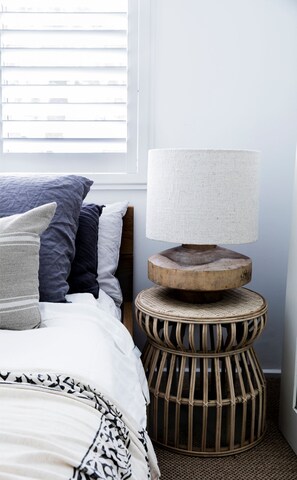 Un lit bien fait, avec une couverture à motifs et des oreillers, un tabouret rond en bois avec un siège en osier, et une lampe fixée au mur.