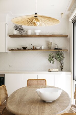 Une cuisine moderne avec des meubles blancs, des étagères en bois et une table de salle à manger ronde entourée de chaises en osier.