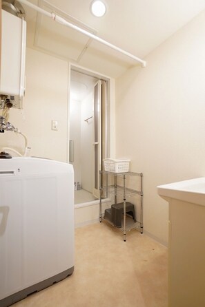 A compact bathroom with a white sink, a large white washing machine, a metal shelf with a basket, and a shower area. - IK Minami6Jo Residence 1003 (Sapporo)