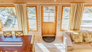 Een gezellig interieur van een chalet met een houten eettafel, een bank met decoratieve kussens en grote ramen met gordijnen.