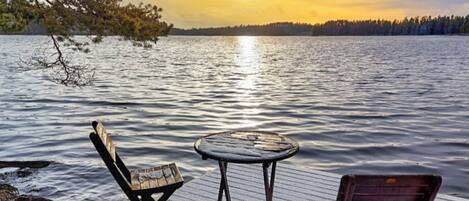 Une terrasse en bois avec deux chaises et une petite table ronde, d’où l’on peut admirer un lac paisible au coucher du soleil.
