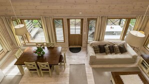 Interieur van een houten chalet met een eettafel, stoelen, een bank en grote ramen met uitzicht op een terras en bomen.