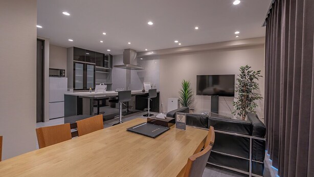 A modern kitchen with a dining area, including a wooden table and chairs, a small refrigerator, and a flat-screen TV mounted on the wall. - Residence Hotel Takayama Station (Takayama)