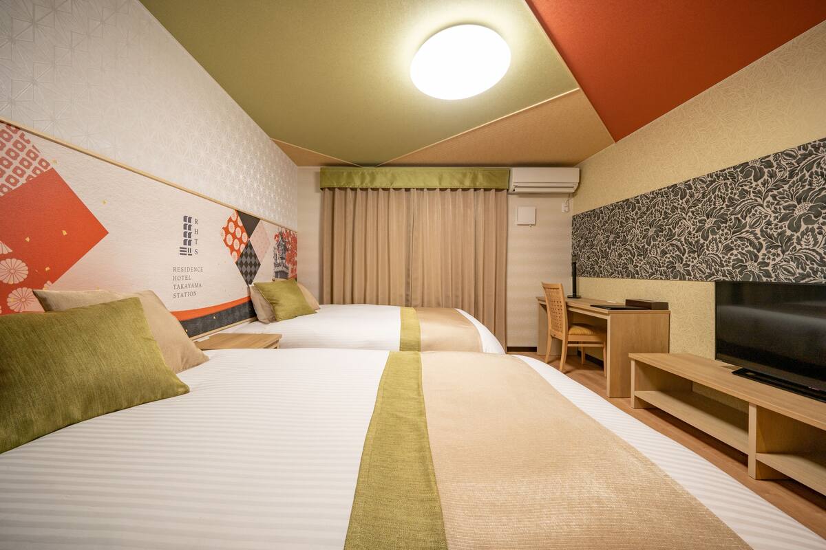 A hotel room with two beds, a desk, a chair, a television, and a wall with a floral pattern.
