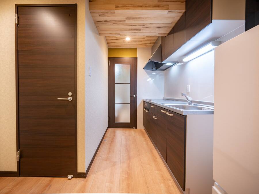 Un pasillo estrecho con suelo de madera, una puerta de madera oscura y una zona de cocina con un lavabo y armarios.