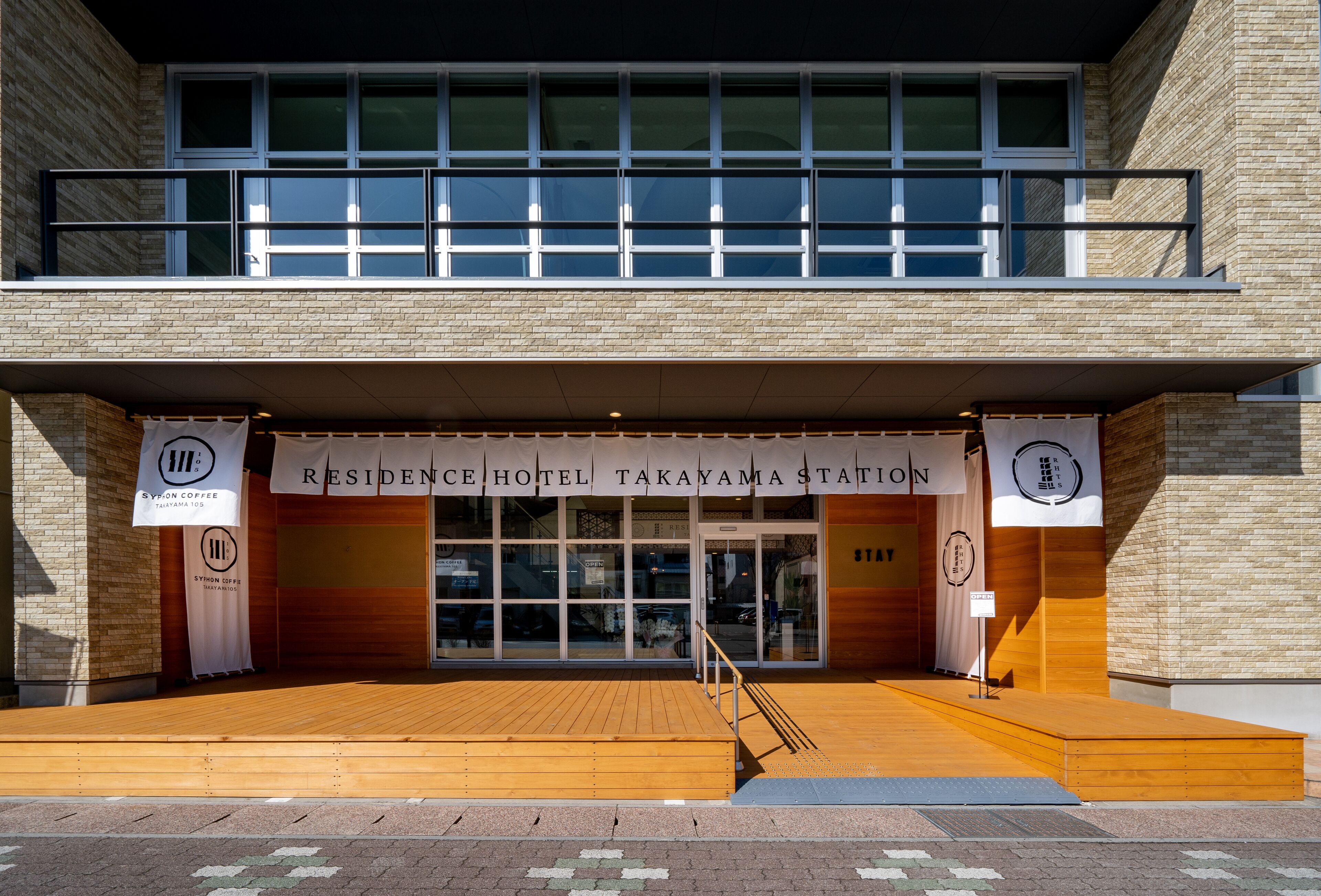 The exterior of the Residence Hotel Takayama Station, featuring a modern design with a wooden deck and clear signage.