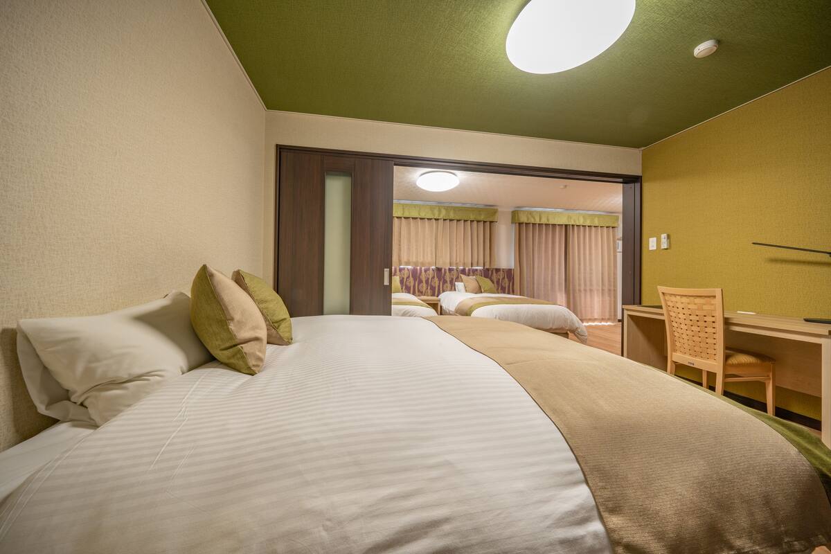 A hotel room with a large bed, a desk, and a chair. There is a door leading to another room.