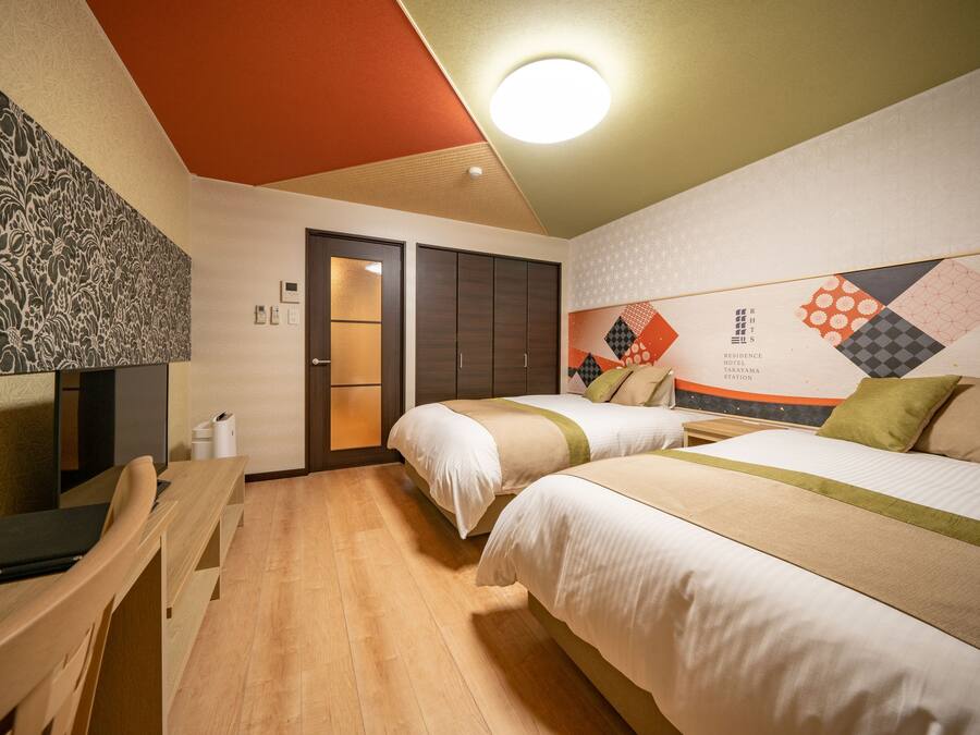 A hotel room with two beds, a desk, and a chair. There is a door leading to a bathroom and a wall with a geometric pattern.