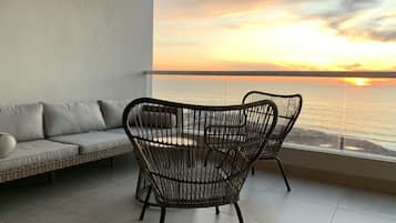A balcony with a wicker chair and a sofa facing a sunset over the ocean.