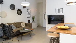 A modern living room with a sofa, coffee table, and a dining area with a bowl of fruit. - Garden 1 Bedroom Apartment (Tel Aviv)