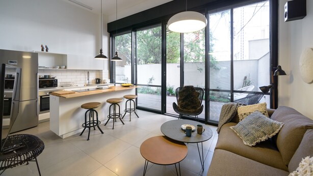 A modern kitchen with a central island, bar stools, and a large window providing a view of the outdoors. - Garden 1 Bedroom Apartment (Tel Aviv)