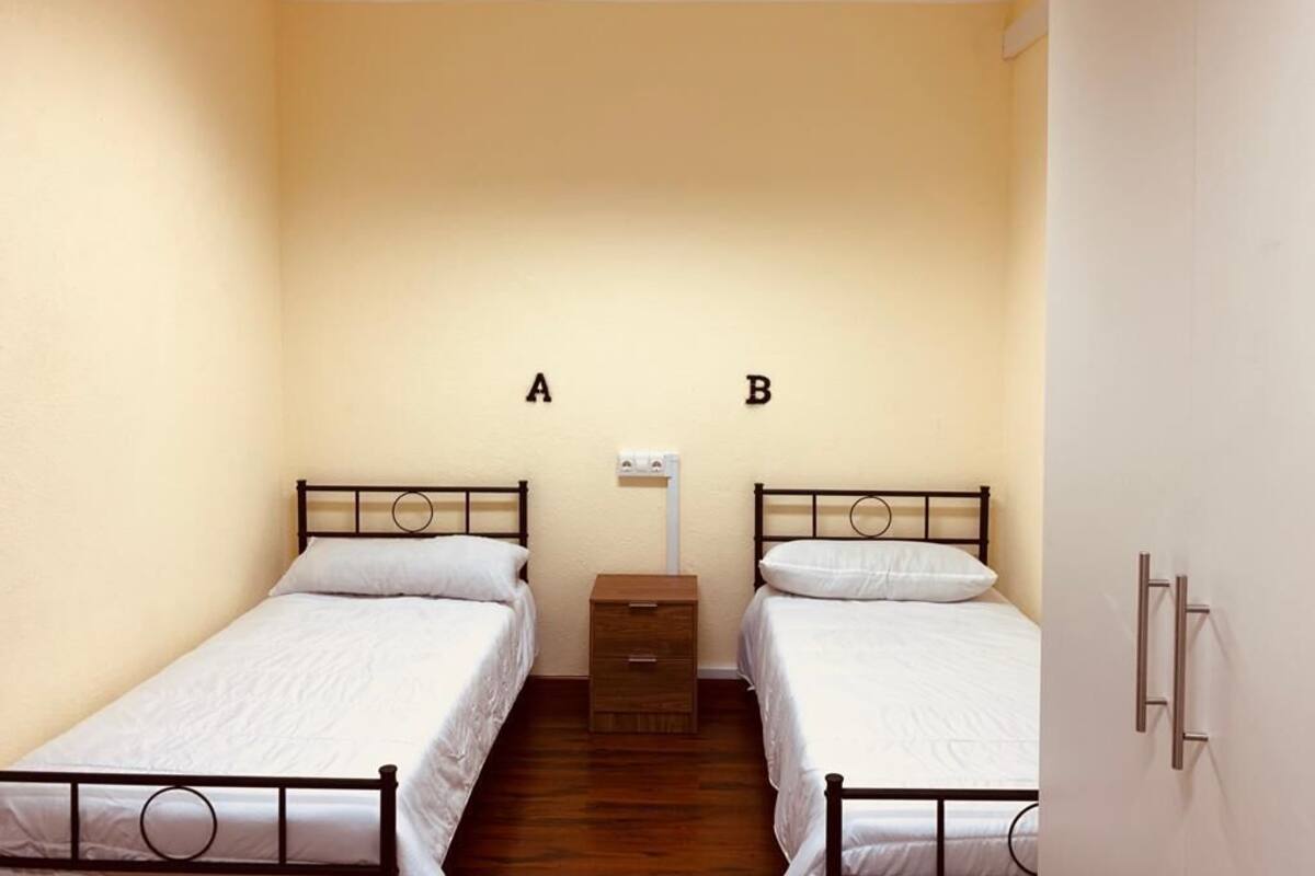 Een kamer met twee eenpersoonsbedden, voorzien van labels A en B, een klein houten nachtkastje en een witte muur met een lichtschakelaar.