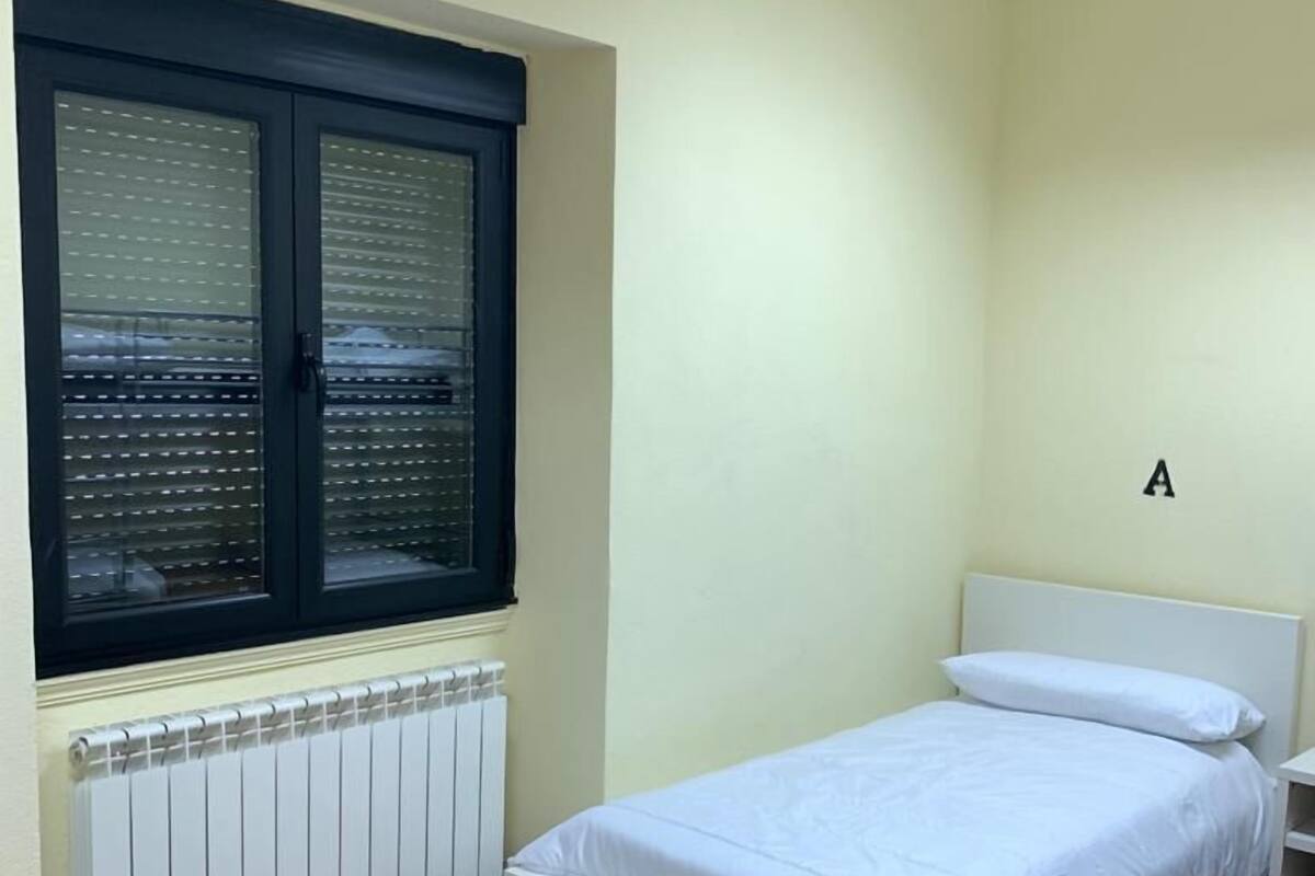 Een slaapkamer met een bed, een raam met zonwering, een radiator en een letter 'A' op de muur.