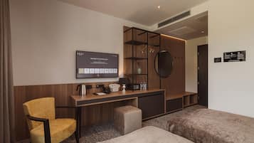 A modern hotel room with a wooden desk, a television, a sofa, and a small ottoman.