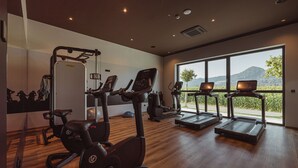 A gym with cardio machines, a weight bench, and a large window with a view of greenery and hills. - Hotel Grof (Vransko)