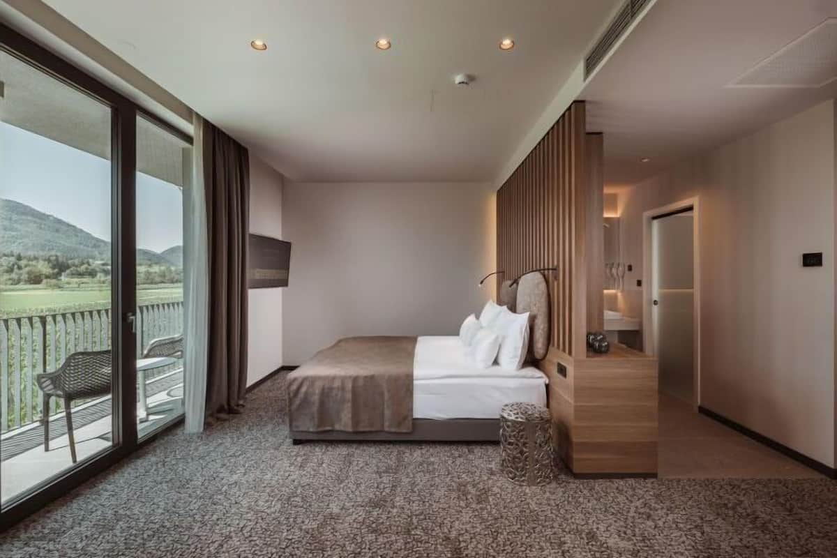 A hotel room with a large bed, a view of the outdoors, a balcony with a chair, and a wooden panel wall.