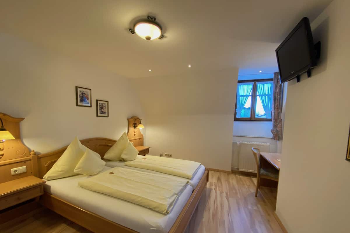 A bedroom with a wooden bed, bedside lamps, a flat-screen TV mounted on the wall, and a window with curtains.