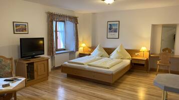 Quarto com cama de madeira, televisão sobre um suporte, janela com cortinas e mesa de cabeceira com abajur.
