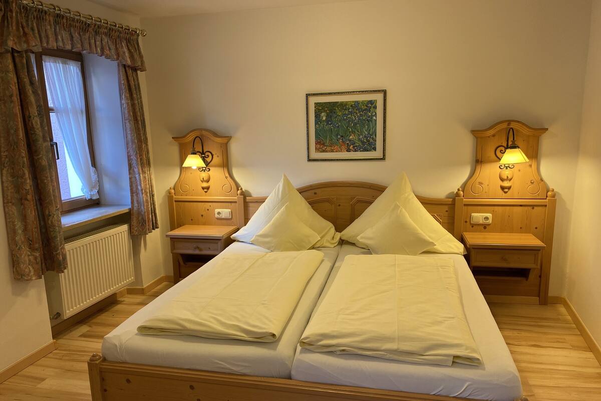 A double bed with two nightstands, each with a lamp, a radiator, and a window with curtains.