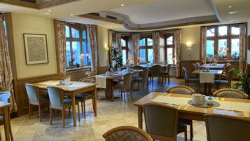 Une salle à manger d’hôtel avec de nombreuses tables et chaises, de grandes fenêtres avec des rideaux et des lampes fixées au mur.