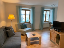 Una sala de estar con sofá, sillón, mesa de centro, televisor y ventanas con cortinas.