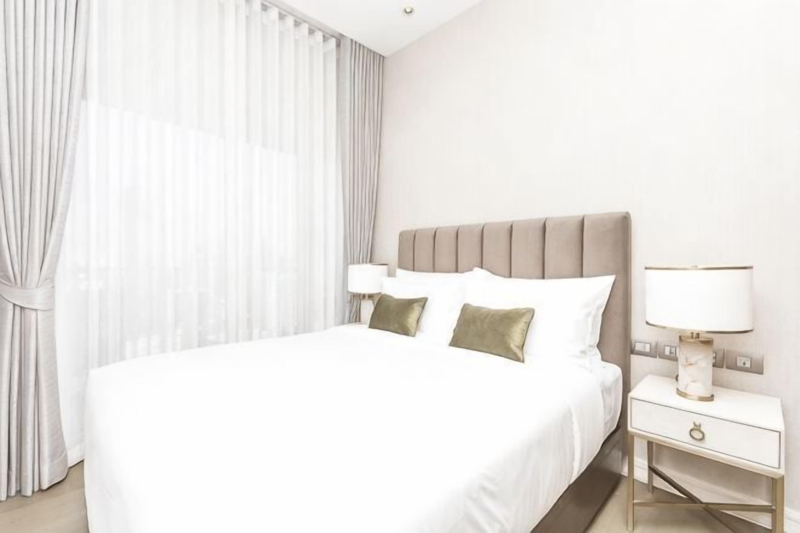 Habitación de hotel con una cama grande, una mesita de noche con lámpara y una ventana con cortinas traslúcidas.