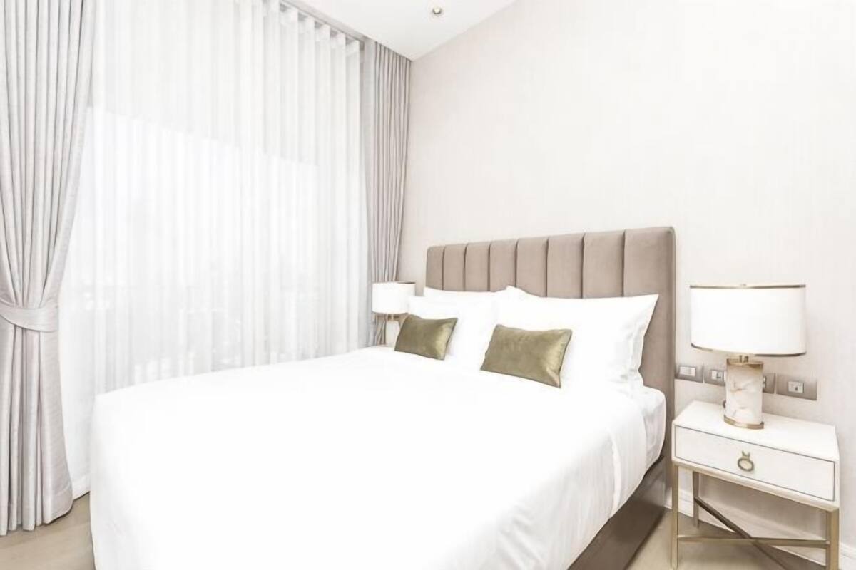 Habitación de hotel con una cama grande, una mesita de noche con lámpara y una ventana con cortinas traslúcidas.